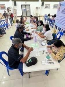 At a long white table, vision screeners are seated on one side and Philippine individuals in need of eyewear are seated on the other. Each vision screener is working with a patient to fill out paperwork and fit their new glasses. 