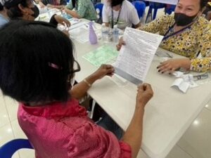 A Philippine woman in a pink shirt reads text on a paper to test her new eyewear. In the background, others are seen also reading and filling out paperwork. 
