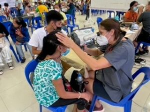 A Mission worker screens a Philippine woman's eyes using a screening device. In the background, many others are also getting their vision screened and fitted for eyewear. 