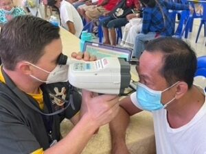 A Mission worker uses a vision screening machine to test a Philippine man's eyesight. They sit across from each other at a table and both are wearing masks. 