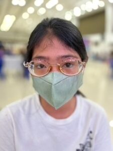 A young Philippine woman in a white shirt and light green mask is photographed with her new pair of glasses, which have circular, gold frames. 