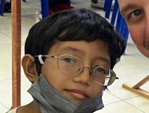 A young Philippine boy poses for a photo in his new eyeglasses, which have a silver, circular frame. His mask if pulled down slightly so you can see his smile. 