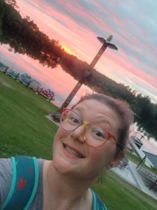 Amanda Dehli, our new Healthcare Supervisor, poses with a smile in front of a sunset over Lions Lake and the Lions Camp totem pole