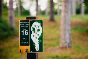 In a wooded area, a Wisconsin Disc Gold Sign for hole #16 is posted with directions around the course. 